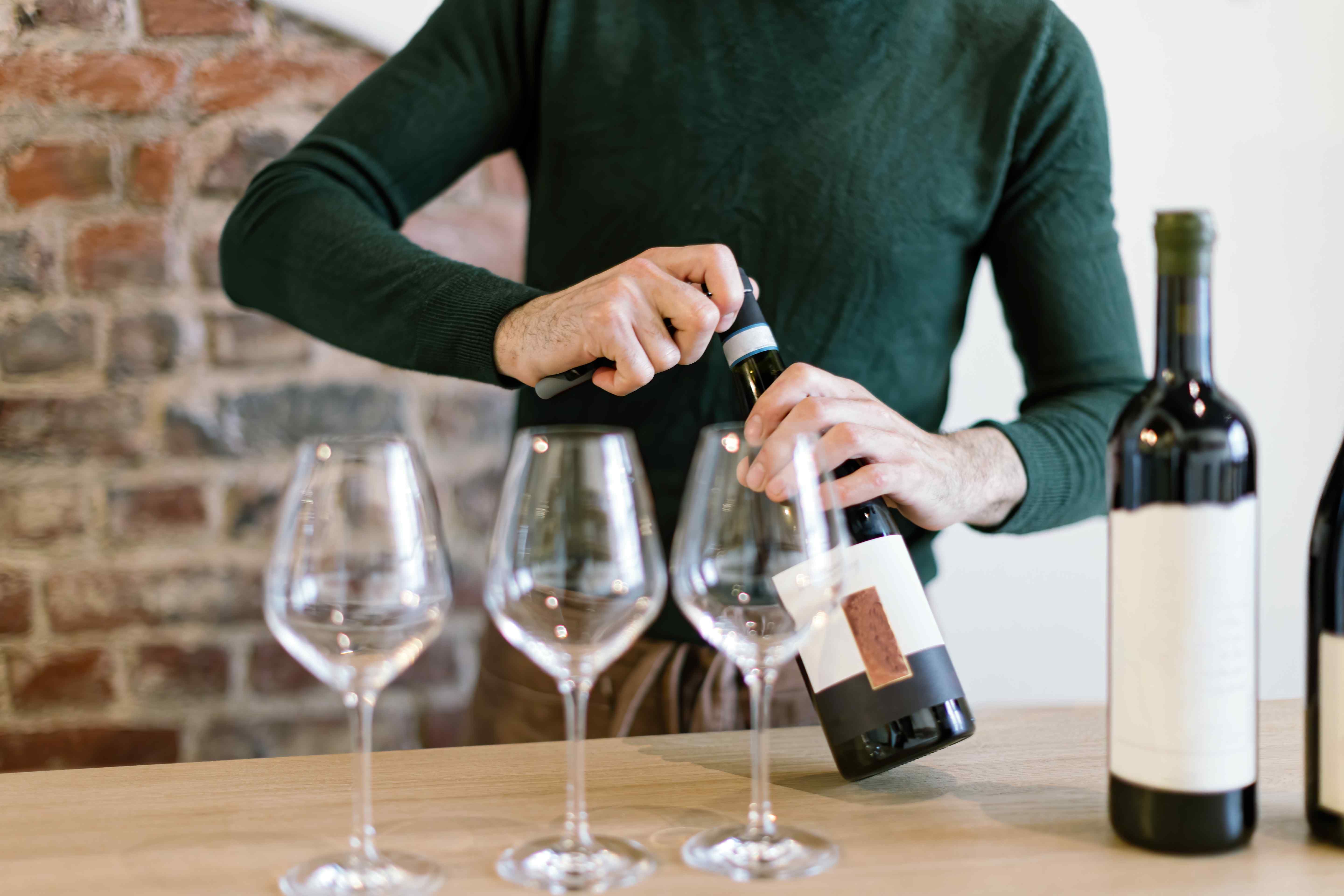 A man standing behind a table, opening a wine bottle to pour into three empty wine glasses.