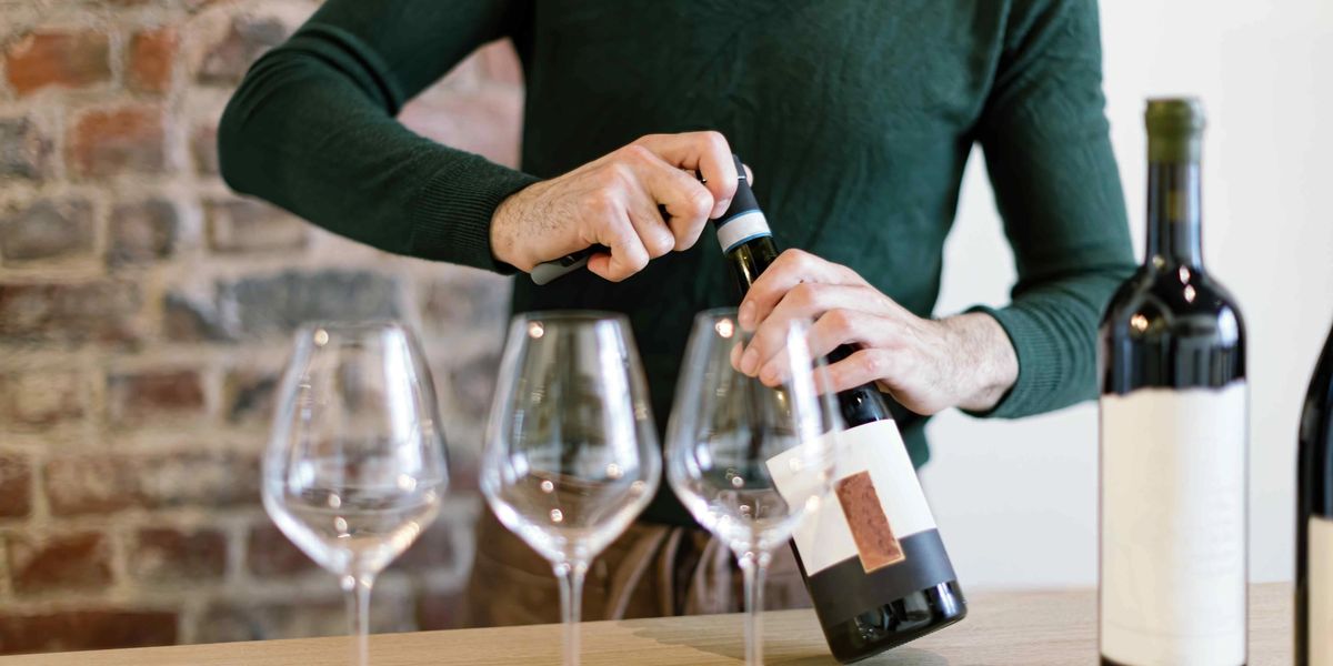 A man standing behind a table, opening a wine bottle to pour into three empty wine glasses.