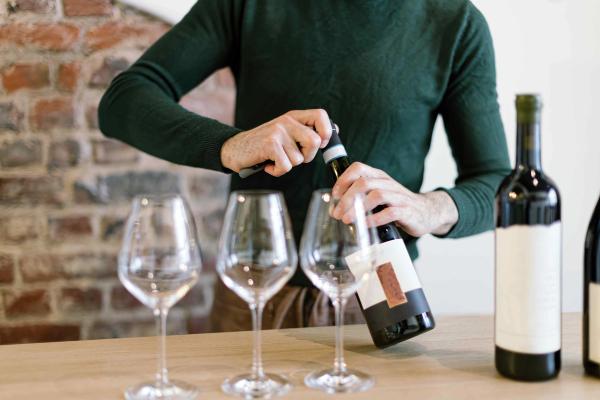 A man standing behind a table, opening a wine bottle to pour into three empty wine glasses.