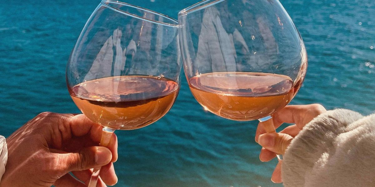 Two people toasting with glasses of rosé wine outdoors by the water on a sunny day.