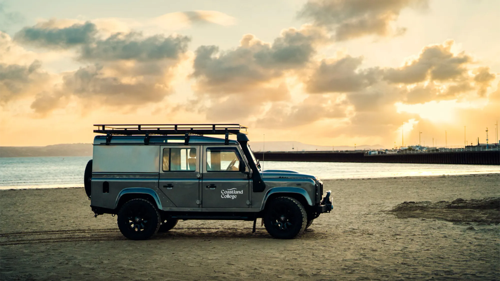 A Coastland College branded Land Rover Defender parked on a beach at sunset