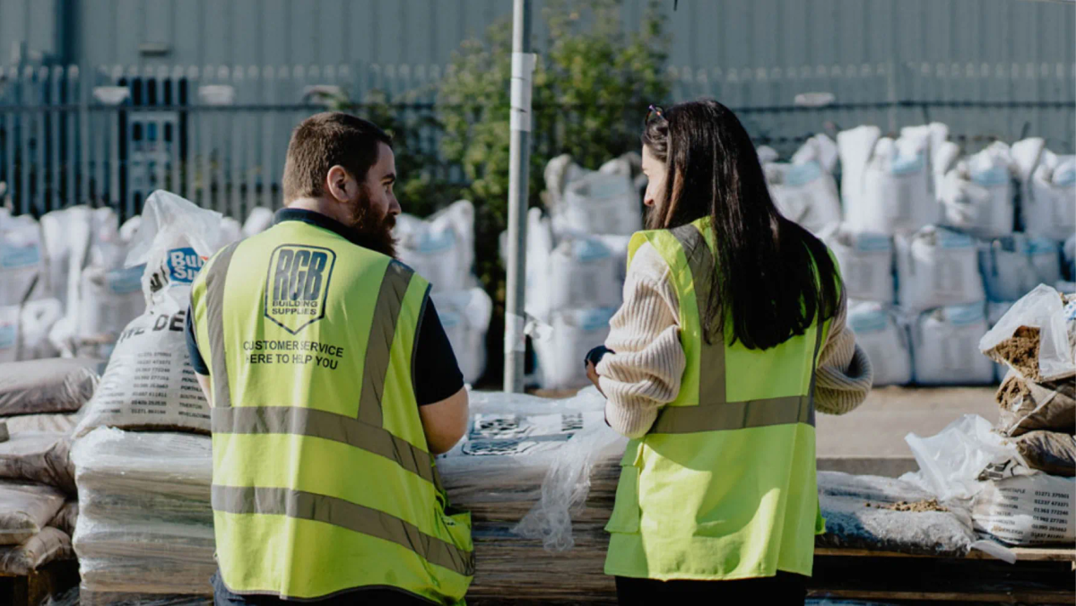 Two RGB Building Supplies staff in high-vis vests standing among stacked building materials in a builders merchant yard