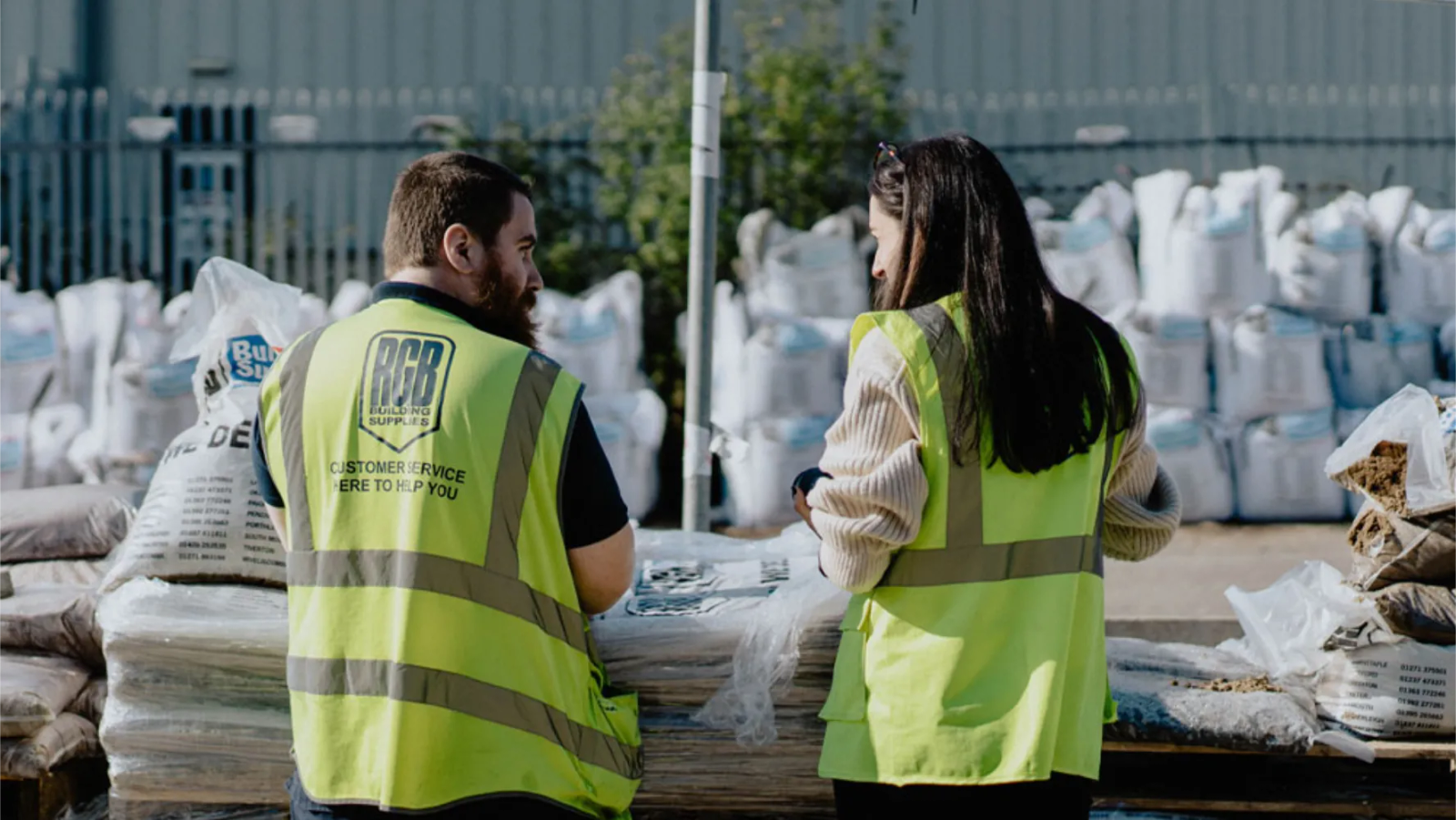 Two RGB Building Supplies staff in high-vis vests standing among stacked building materials in a builders merchant yard