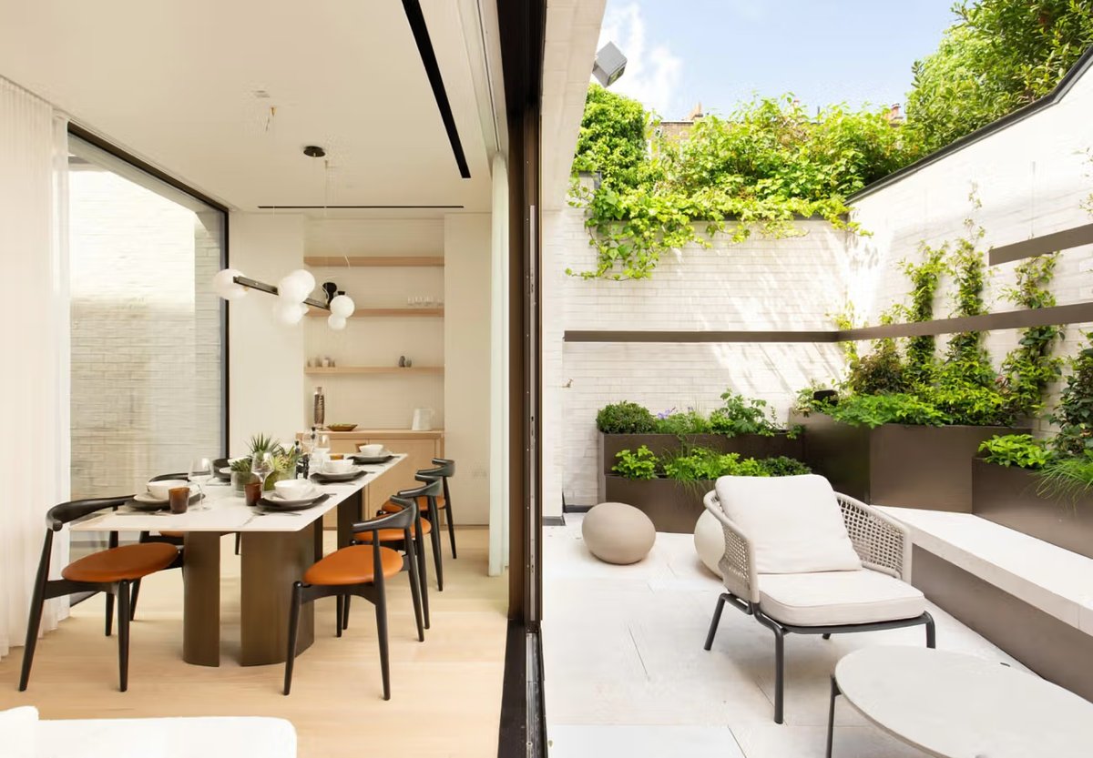 An open-plan dining room with a wooden table and orange chairs leading onto a planted courtyard patio with a lounge chair