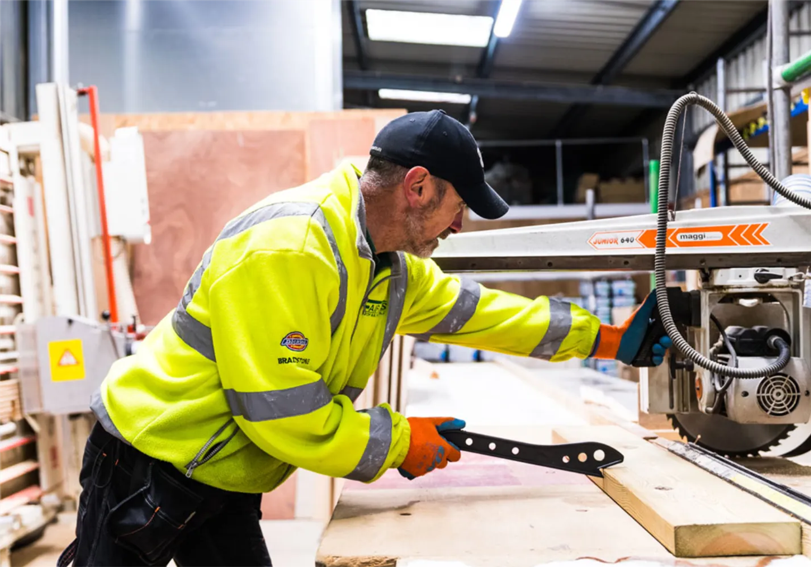A yard worker in high-vis using a radial arm saw to cut a length of timber in a builders merchant yard
