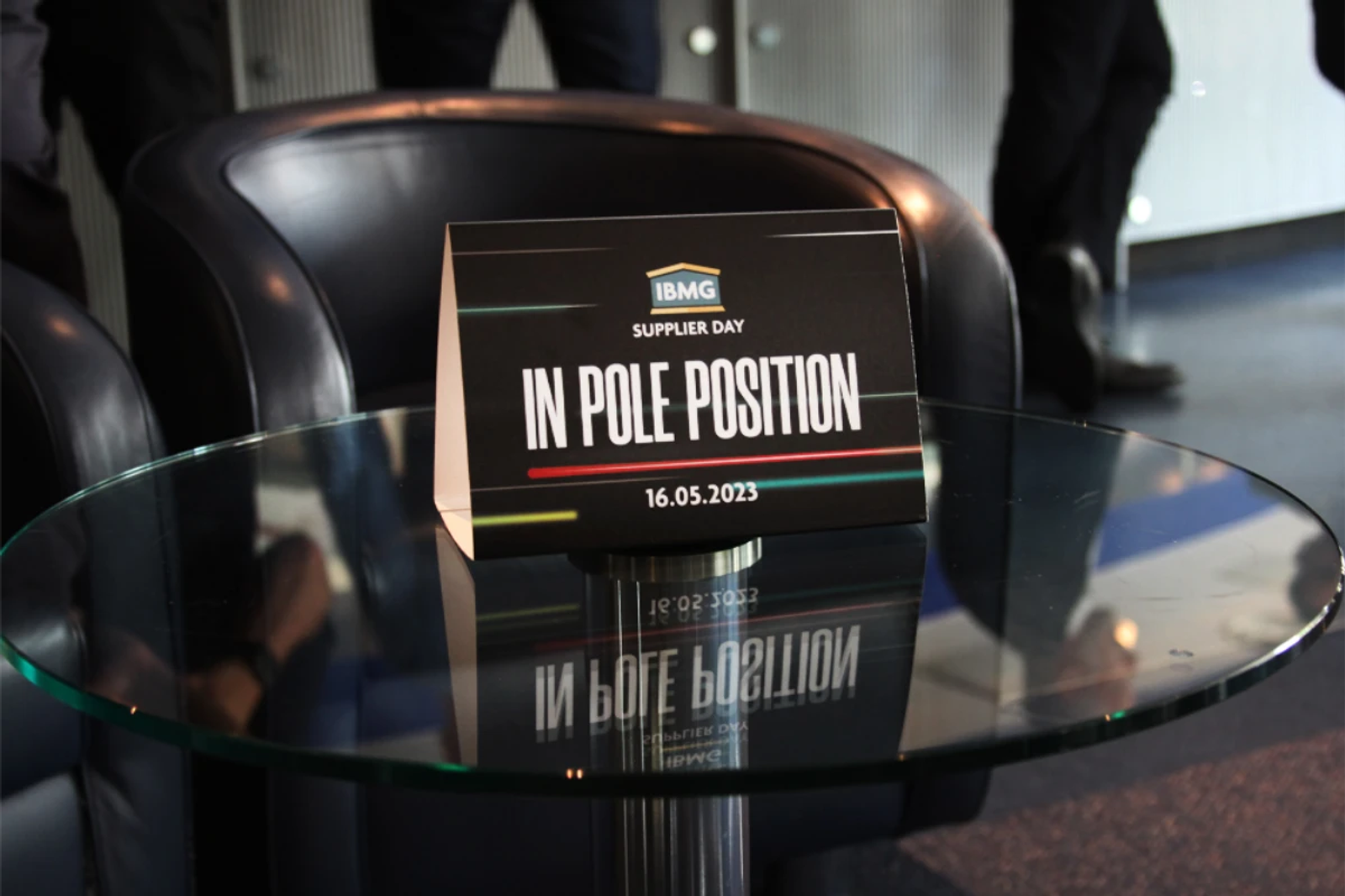 Black table sign reading "IBMG Supplier Day, In Pole Position, 16.05.2023" on a glass table in a lounge area