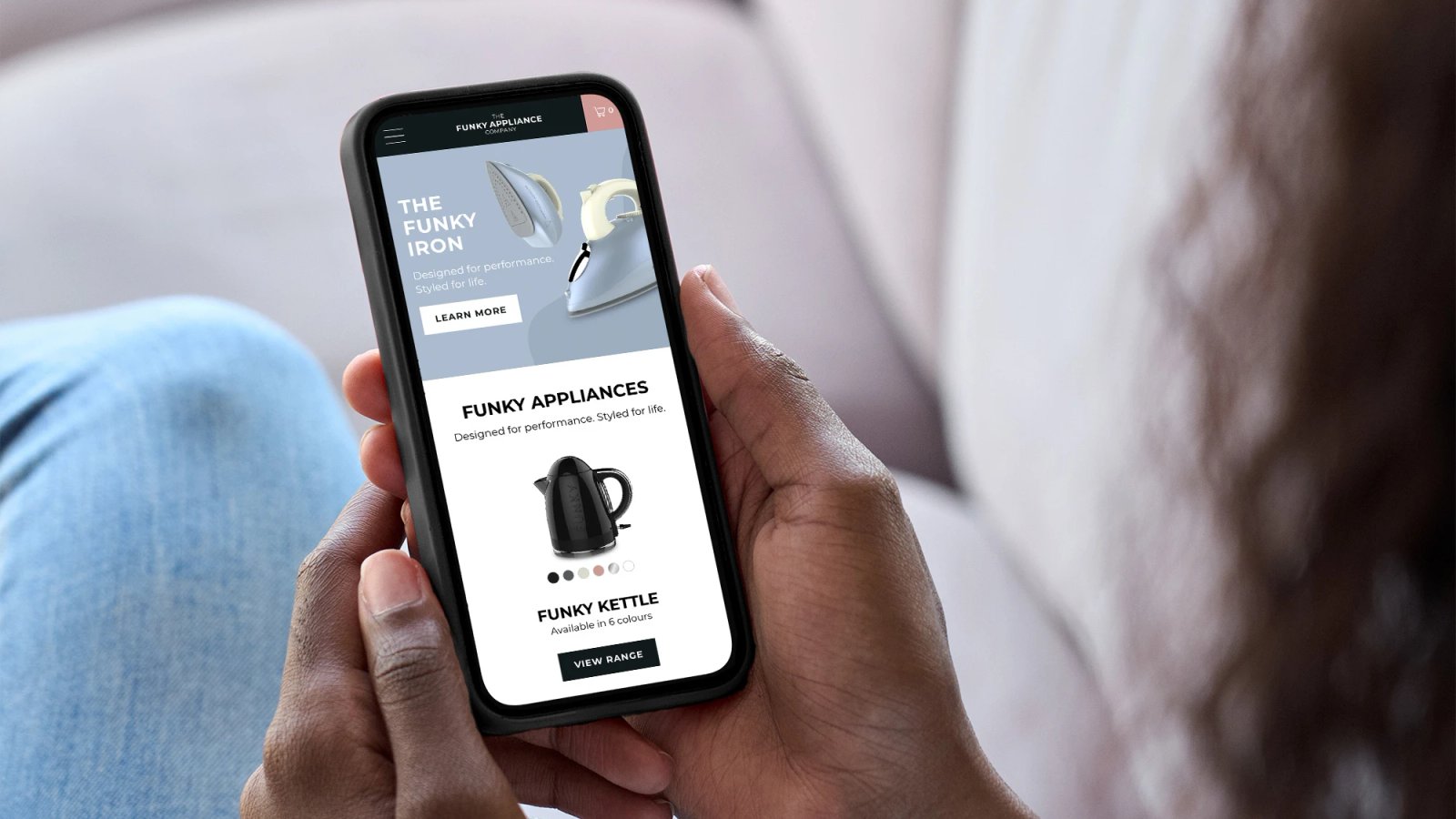 A person holding a phone displaying The Funky Appliance Company website, showing an iron banner and a kettle product page