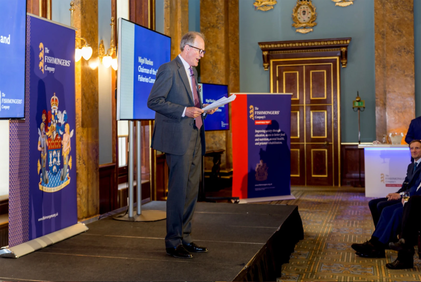 A man in a suit speaking on a stage in front of Fishmongers' Company branded pull-up banners inside a grand panelled hall