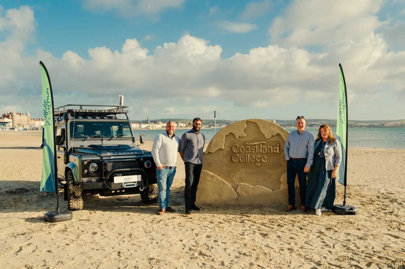 A sand sculpture engraved with the Coastland College name, with a branded Land Rover Defender on the beach