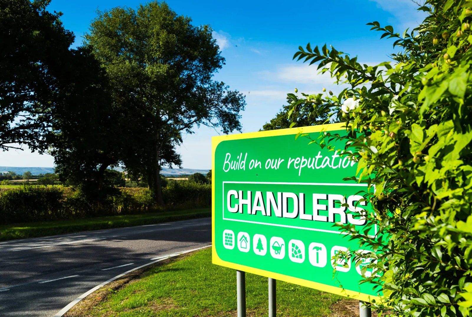 A green Chandlers roadside sign with the 'Build on our reputation' tagline and category icons, set against trees and countryside