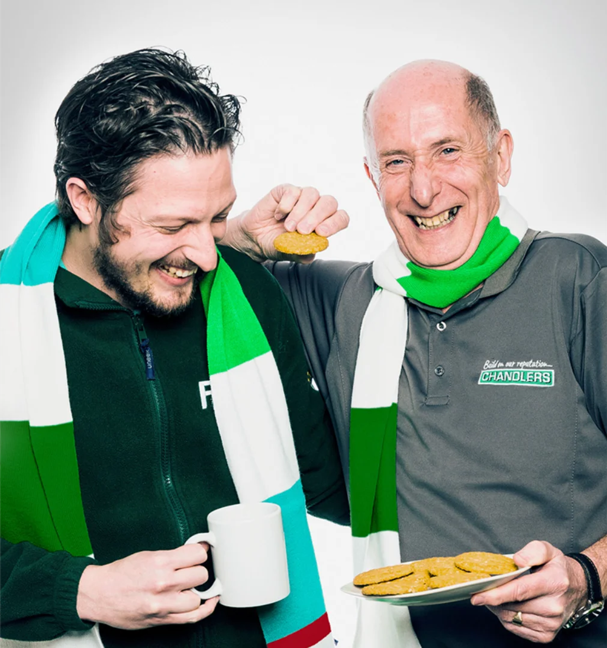 Two staff members wearing green and white scarves, one holding a biscuit and a mug, the other holding a plate of biscuits and wearing a Chandlers polo shirt