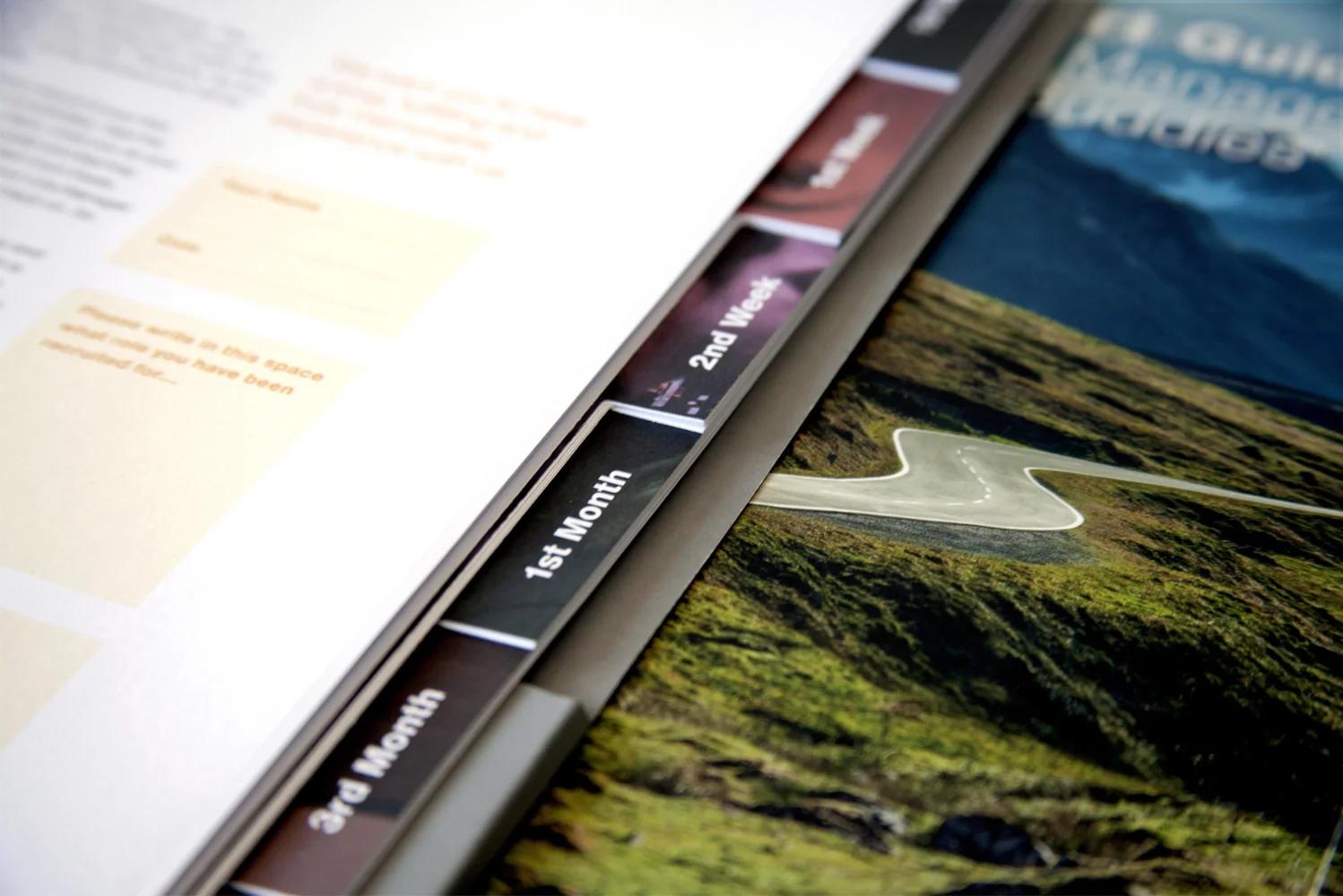 Close-up of road-themed tabbed dividers in the Harwoods Group induction journey booklet, labelled 2nd Week, 1st Month and 3rd Month