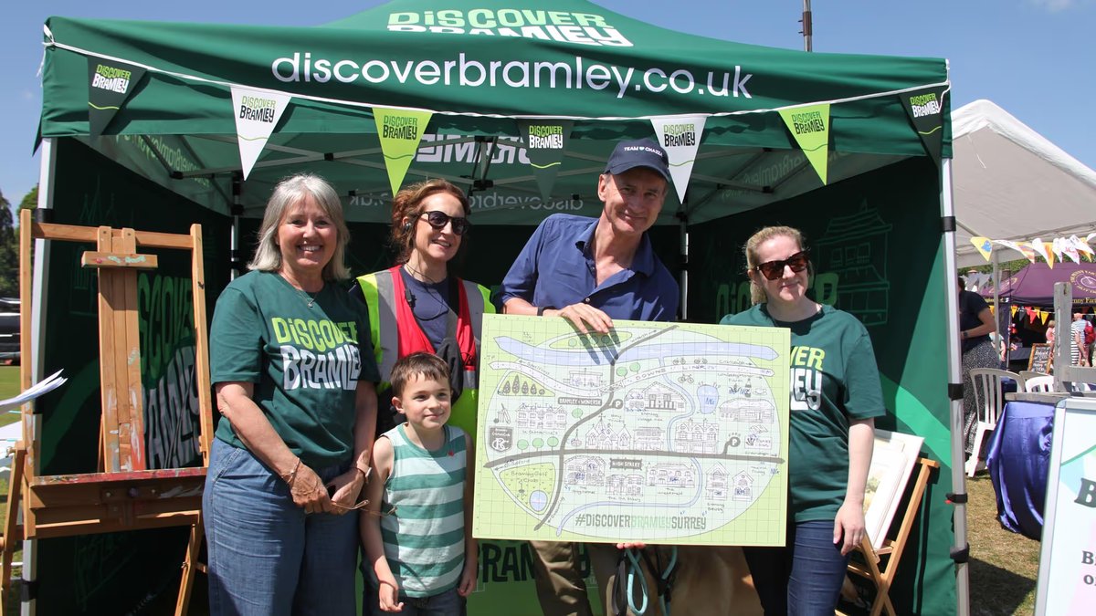 Five volunteers and a child posing under the Discover Bramley gazebo at a summer event, with one man holding an illustrated village map