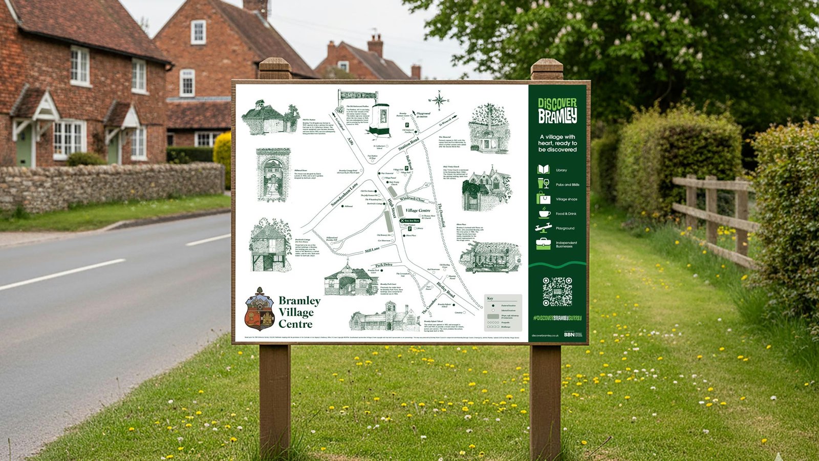 Wooden roadside sign displaying an illustrated map of Bramley Village Centre with a key, local landmarks, and the Discover Bramley logo, standing on a grass verge beside cottages