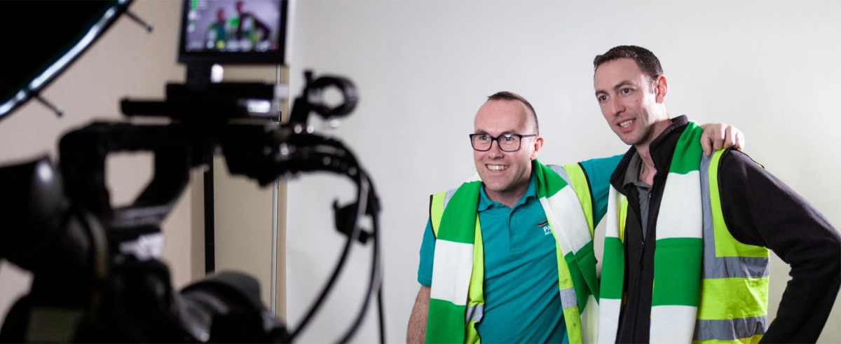 Two staff members wearing green and white scarves over hi-vis vests, smiling next to studio camera equipment