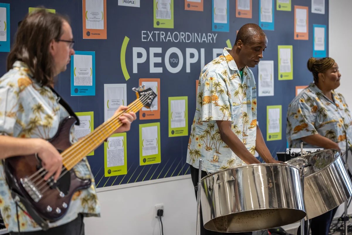A steel band wearing Hawaiian shirts performing on steel drums in front of an Extraordinary People branded wall display