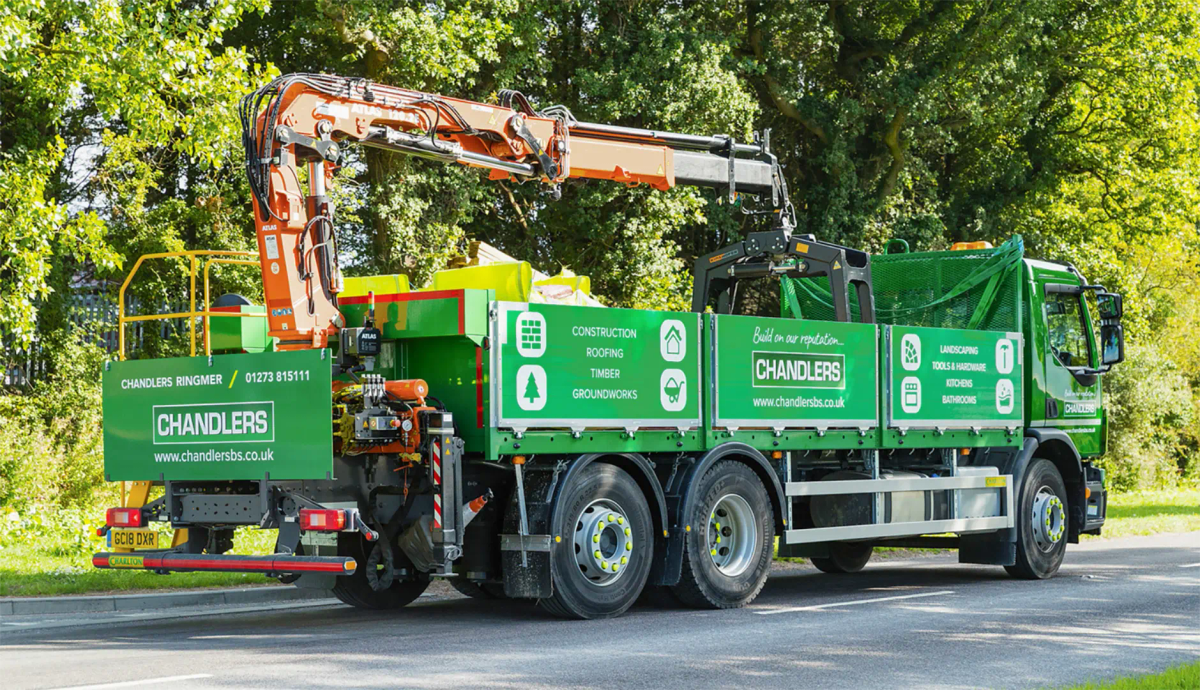 A green Chandlers grab lorry with its crane arm extended, parked beside a roadside Chandlers sign and trees