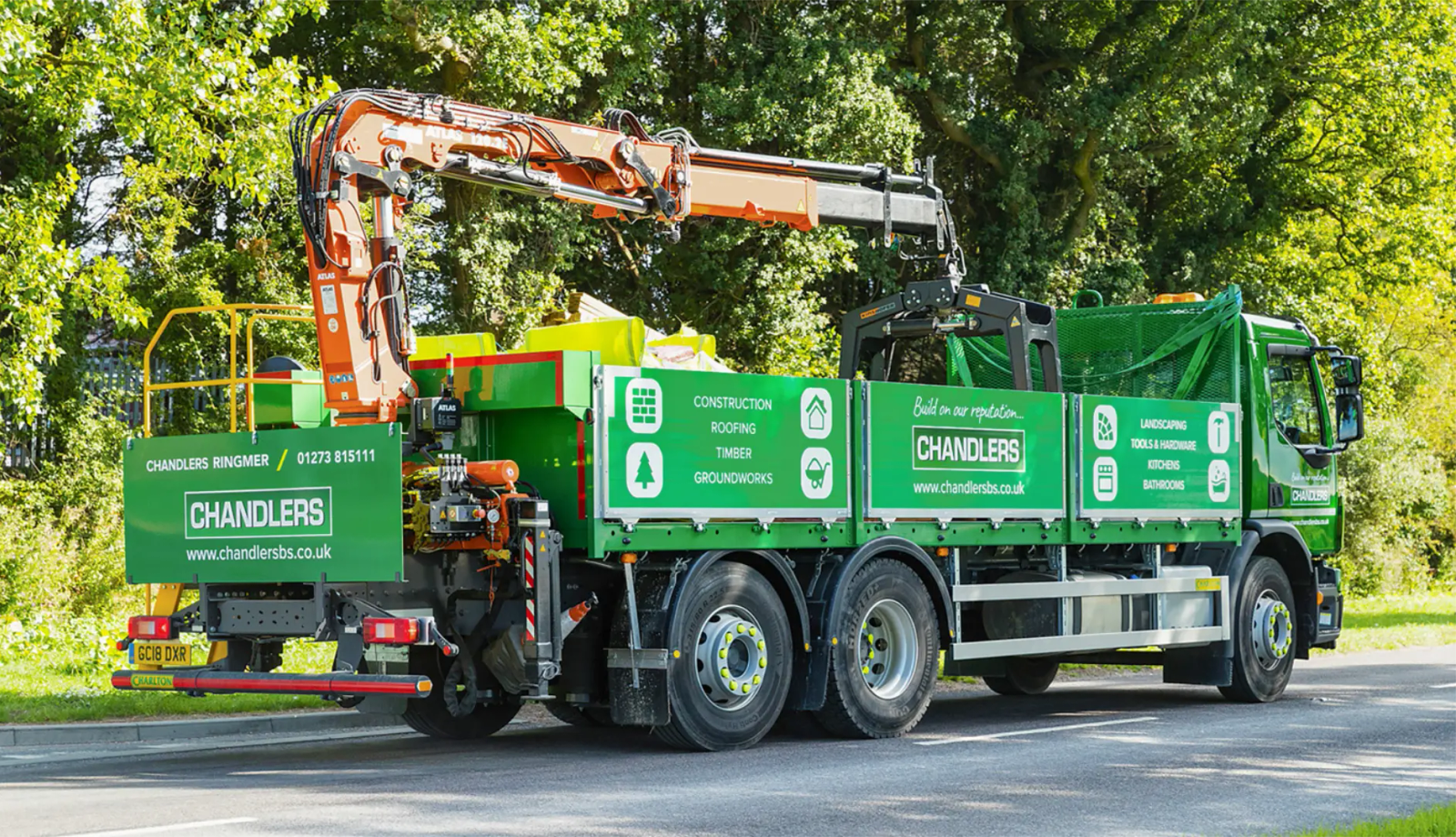 A green Chandlers grab lorry with its crane arm extended, parked beside a roadside Chandlers sign and trees