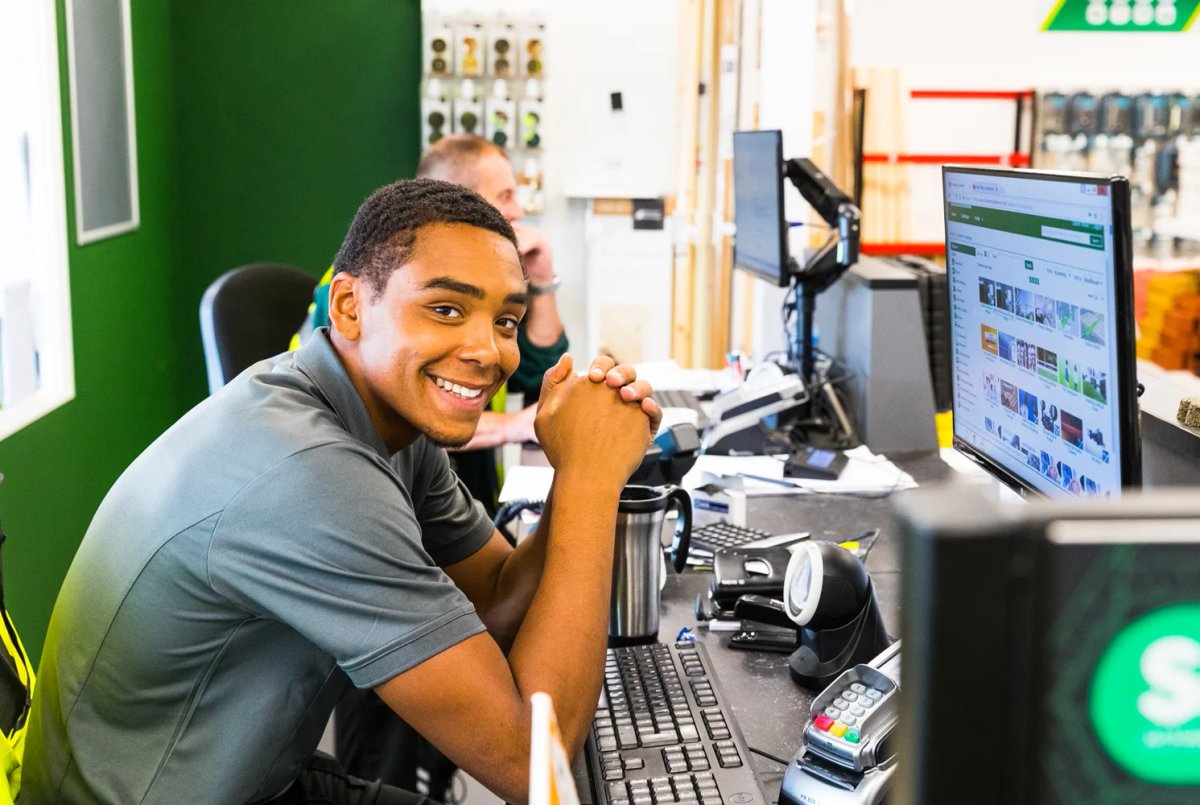 A smiling Chandlers employee sat at a trade counter desk with computer screens, with a colleague in the background