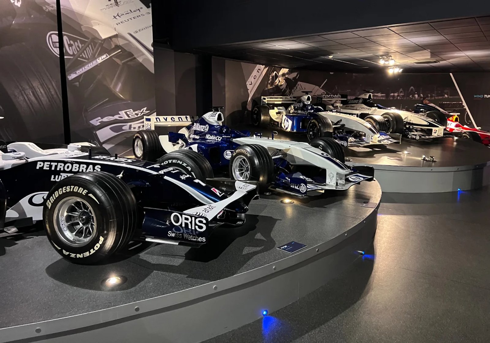 Row of Formula 1 race cars on display, including Williams cars with Petrobras, Allianz and Oris branding