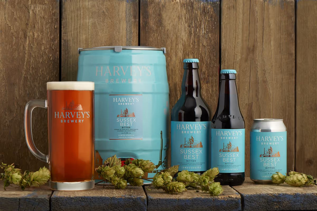 A keg, a stein, two bottles and a can of Harvey's bitter, on a wooden shelf with a wooden wall in the background