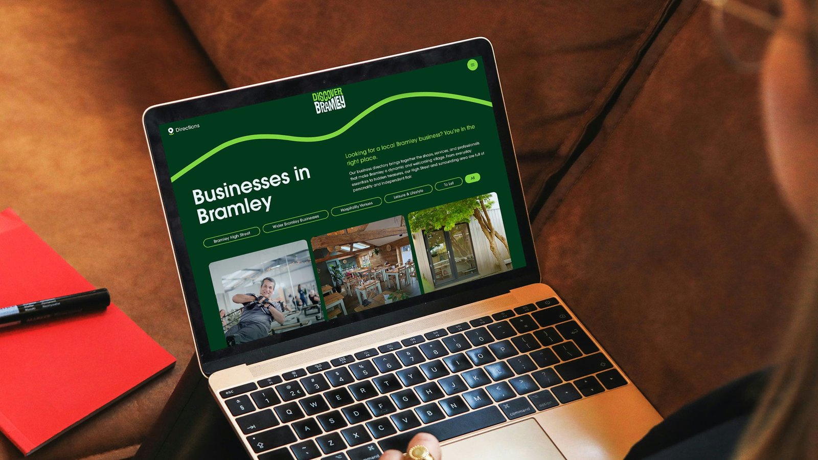 Laptop on a leather sofa displaying the Discover Bramley website 'Businesses in Bramley' directory page, with a red notebook and marker pen beside it