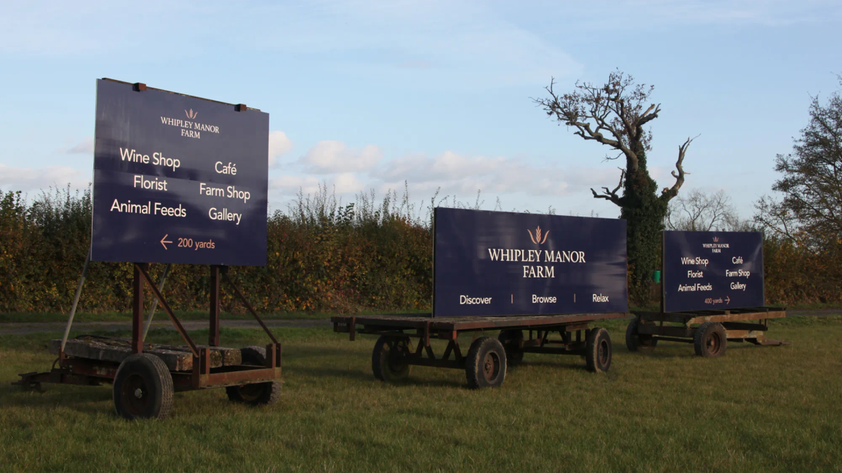 Three large blue Whipley Manor Farm signs on trailers in a field listing wine shop, florist, café, farm shop and gallery.