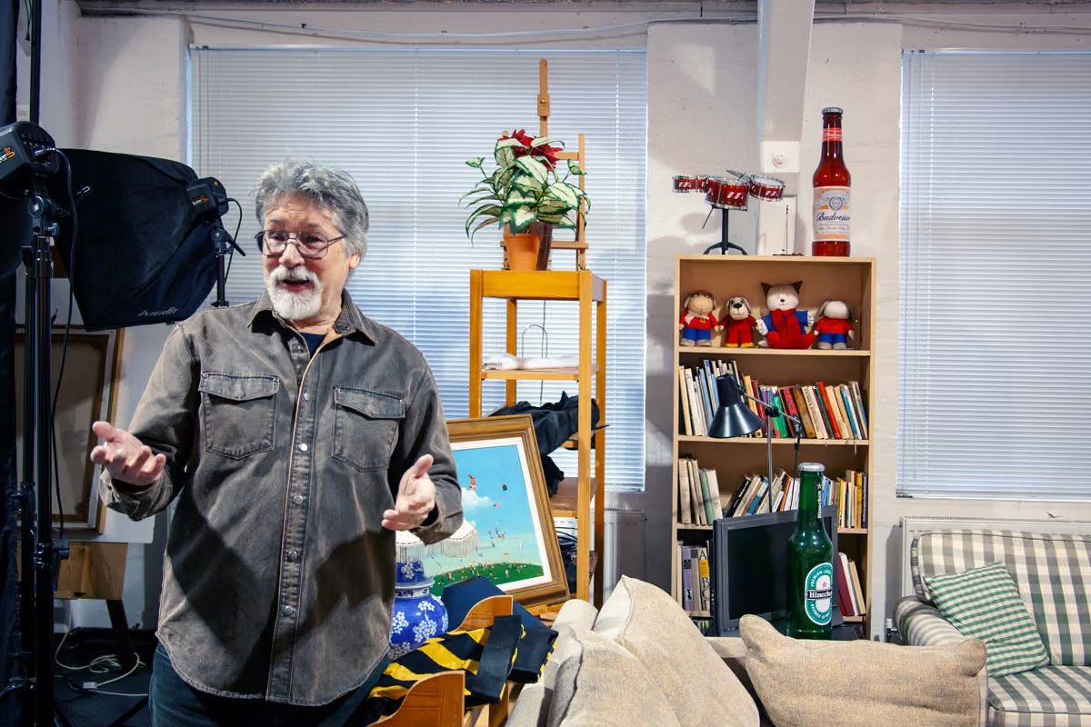 Alex Brychta talking with two colleagues in a studio surrounded by framed paintings and artwork