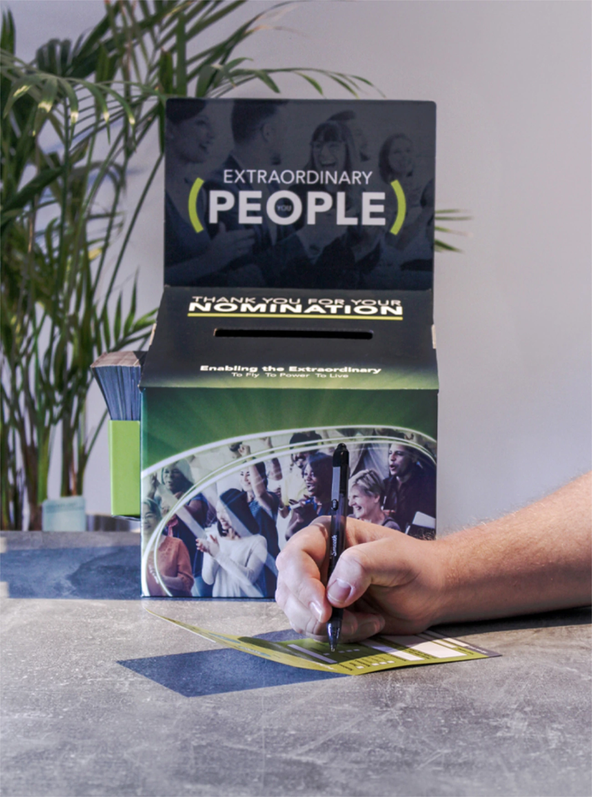 A hand filling in an Extraordinary People nomination form in front of a branded cardboard nomination box