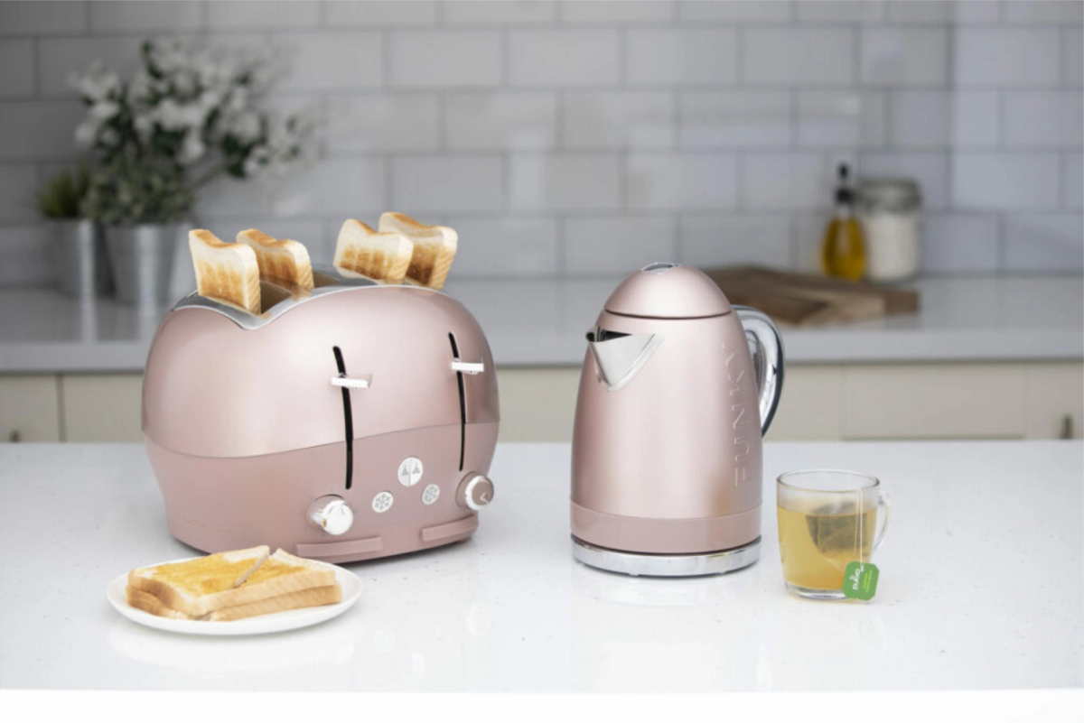 A rose gold kettle and four-slice toaster with toast on a kitchen countertop, beside a cup of tea