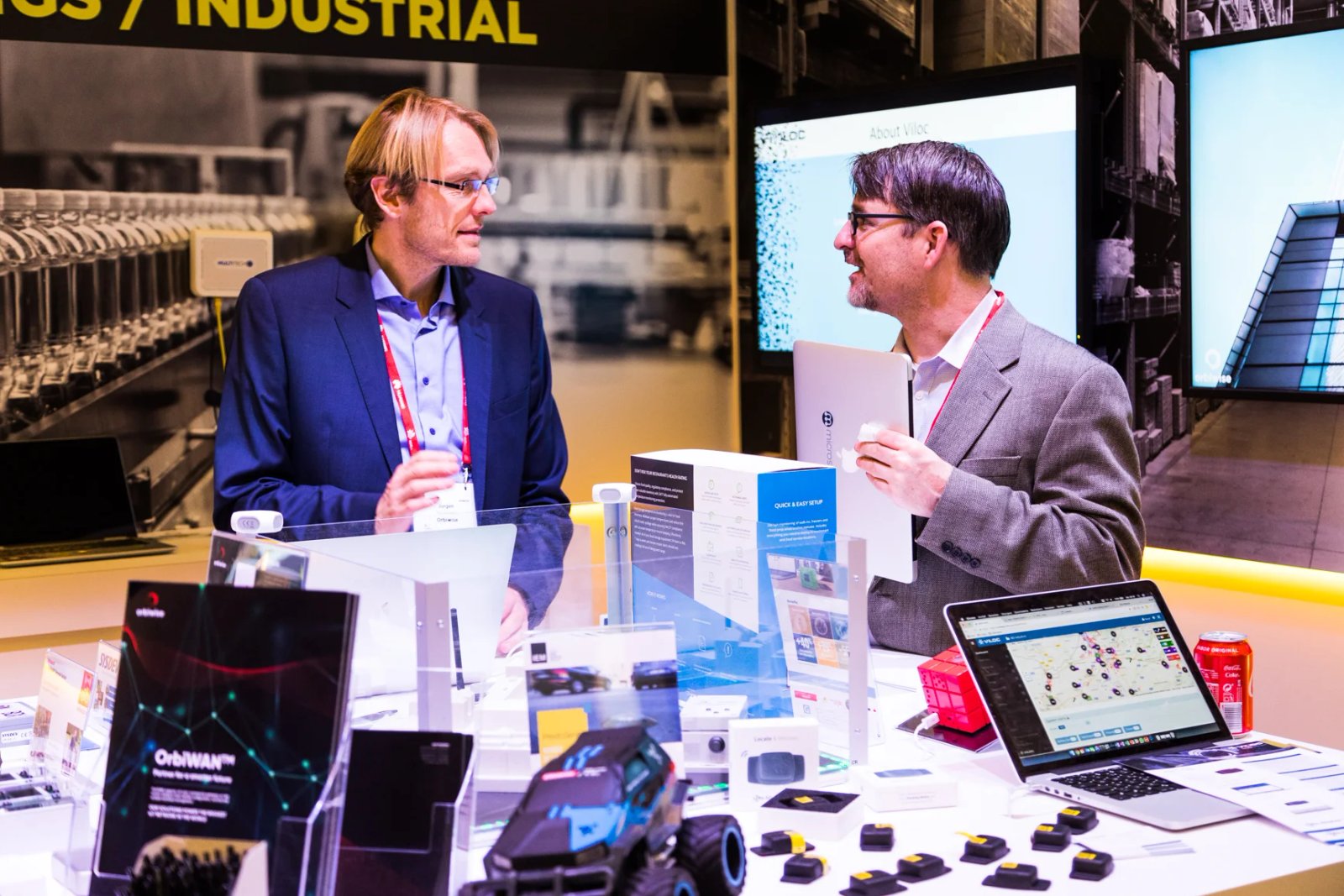 Two men talking at a trade show stand displaying IoT hardware, with laptops and screens showing Orbiwise and Viloc software in the background