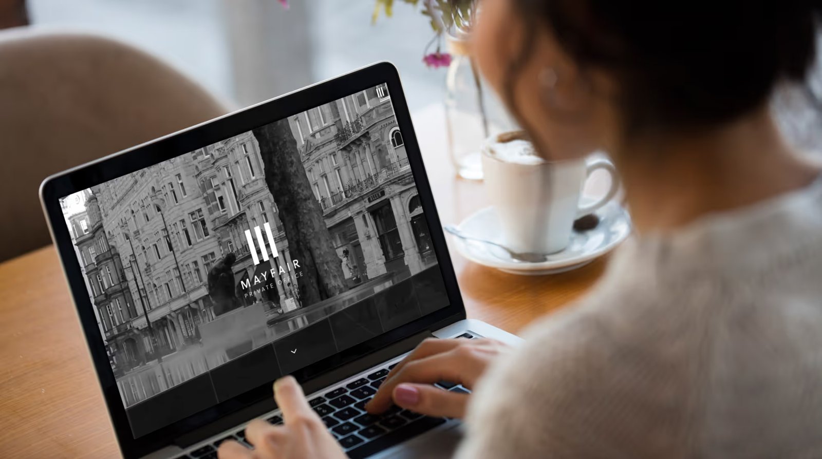 A woman working on a laptop in a café, with the Mayfair Private Office homepage open on screen