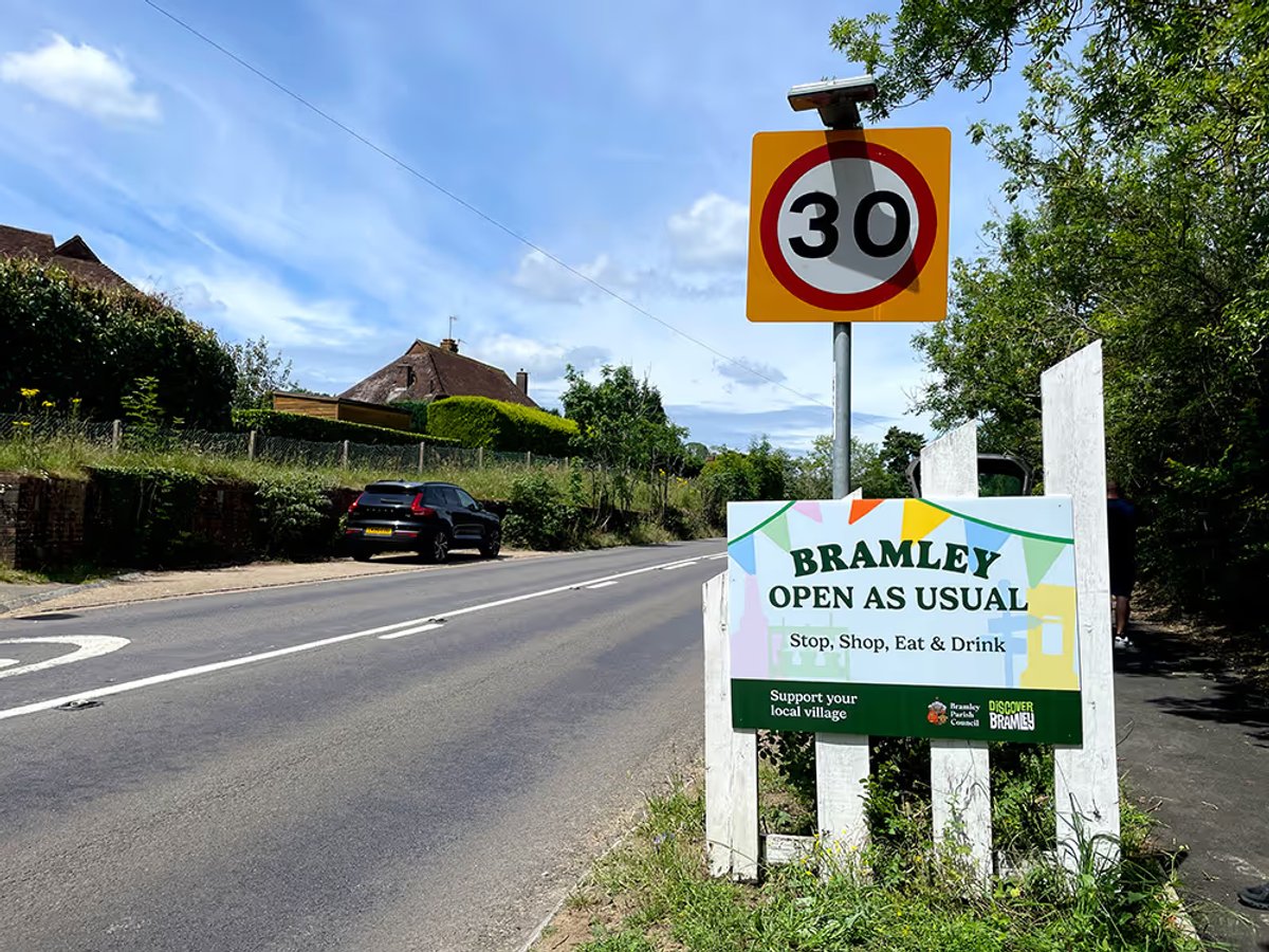 Roadside sign reading 'Bramley Open As Usual - Stop, Shop, Eat & Drink' beneath a 30mph speed limit sign, with a village lane and cottage in the background