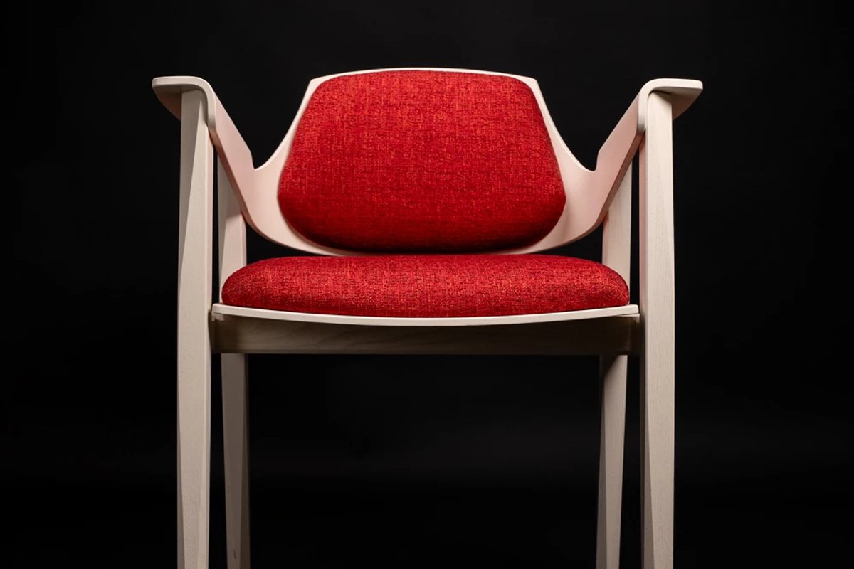 A handcrafted dining chair with a cream wood frame and red upholstered seat, on a black background