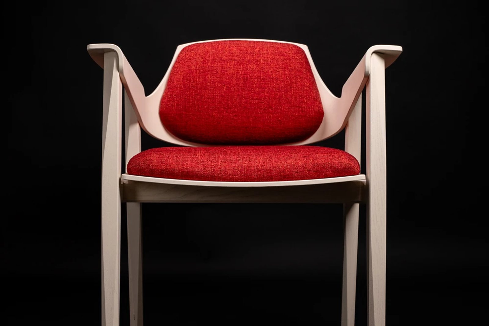 A handcrafted dining chair with a cream wood frame and red upholstered seat, on a black background