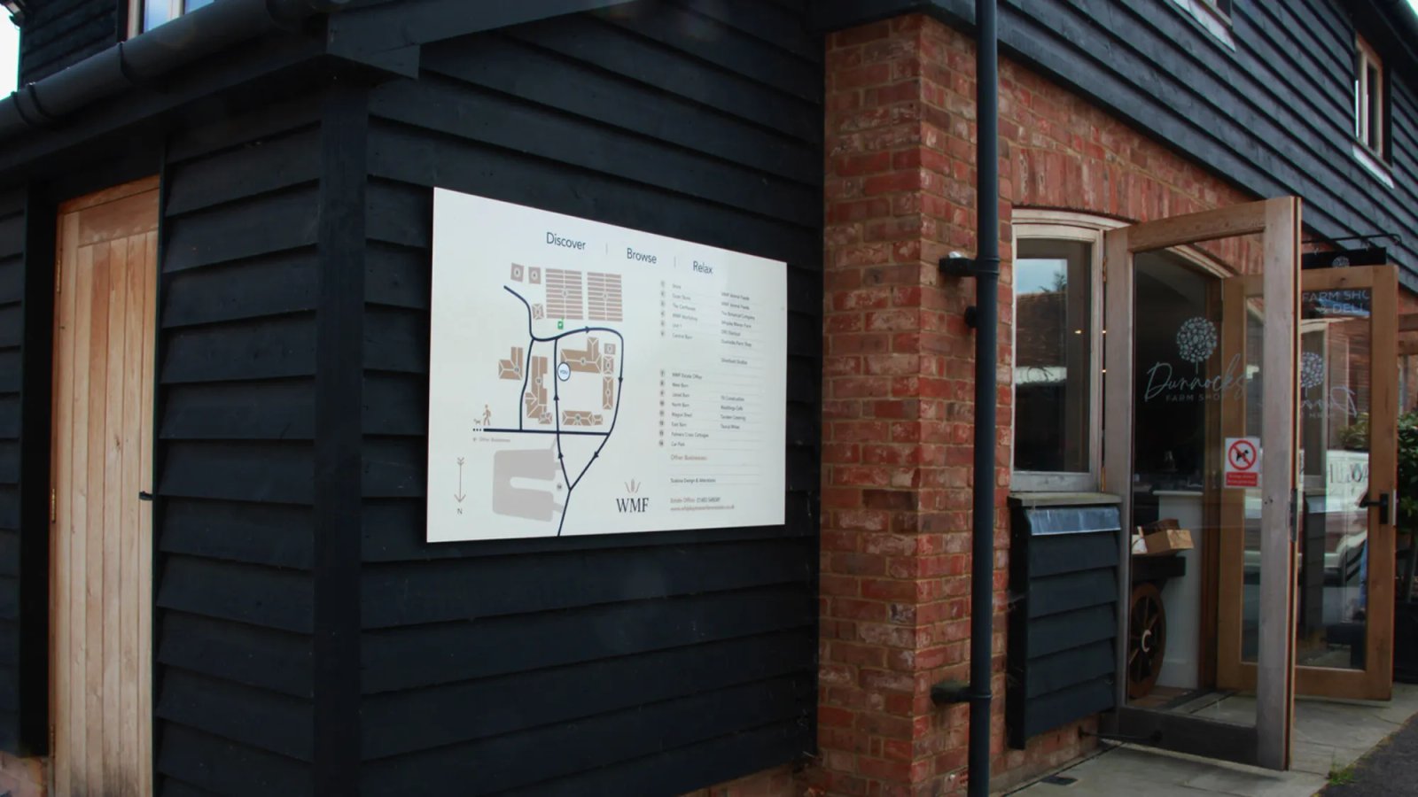 A white site map sign on a black wooden wall beside the brick entrance to Dunnocks Farm Shop at Whipley Manor Farm.