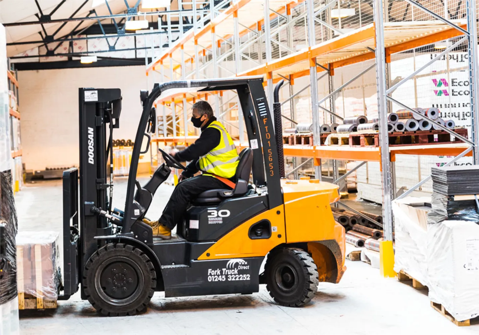 A forklift operator wearing a face mask and hi-vis vest driving a counterbalance forklift through a builders merchant warehouse