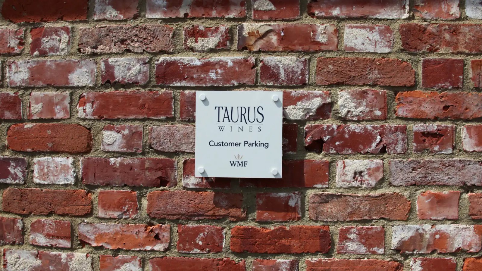 A white Taurus Wines customer parking sign with the WMF logo mounted on a weathered red brick wall.