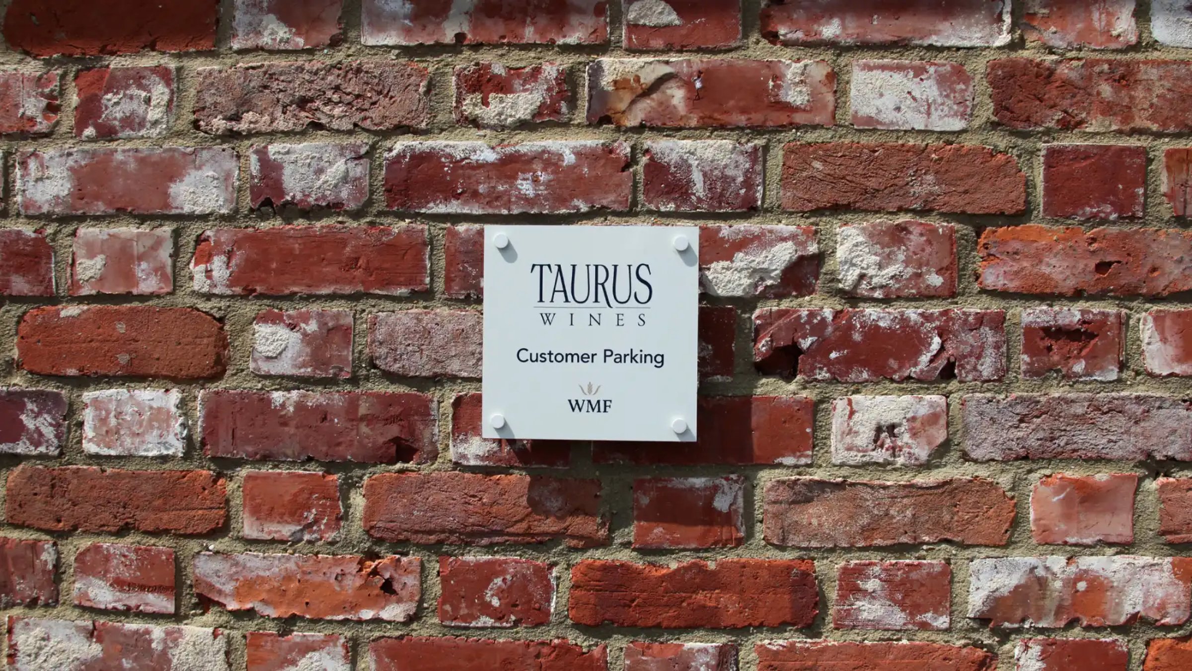 A white Taurus Wines customer parking sign with the WMF logo mounted on a weathered red brick wall.