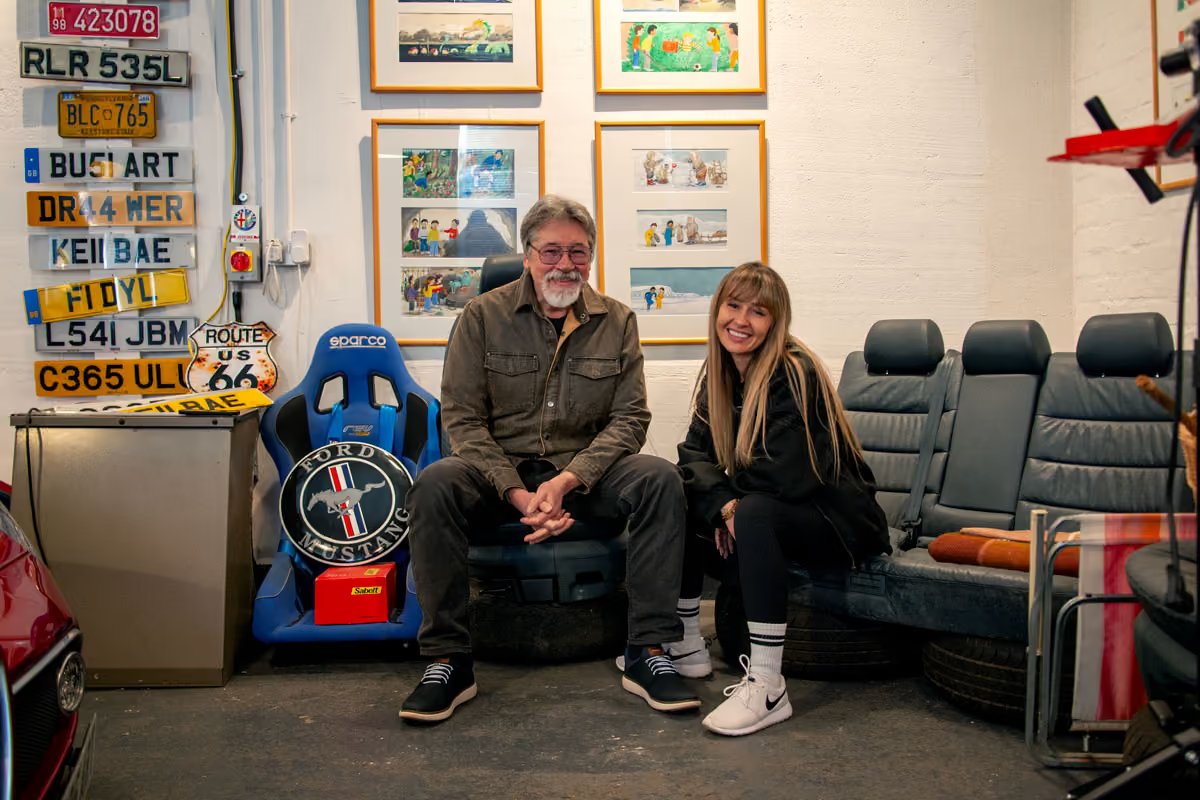 Alex Brychta and Kelli seated together in front of framed illustrations from his children's books