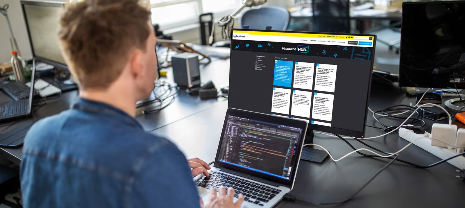A developer coding on a laptop with the LoRa Alliance resource hub website open on a monitor behind