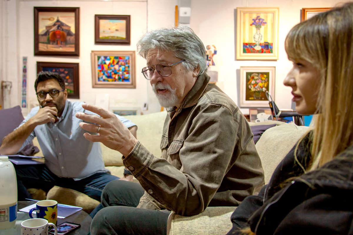 Alex Brychta speaking mid-conversation in his studio, surrounded by original paintings and framed artwork