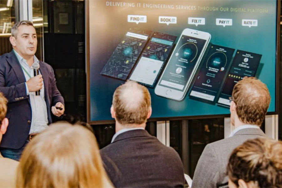 Brett holding a microphone giving a speech with a branded Itarmi slide in the background