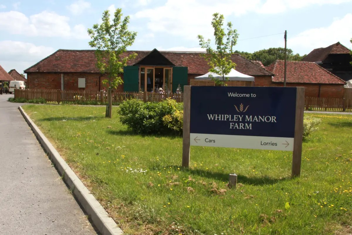 A large welcome sign for Whipley Manor Farm with separate arrows for cars and lorries, set on a grassy lawn in front of brick farm buildings.