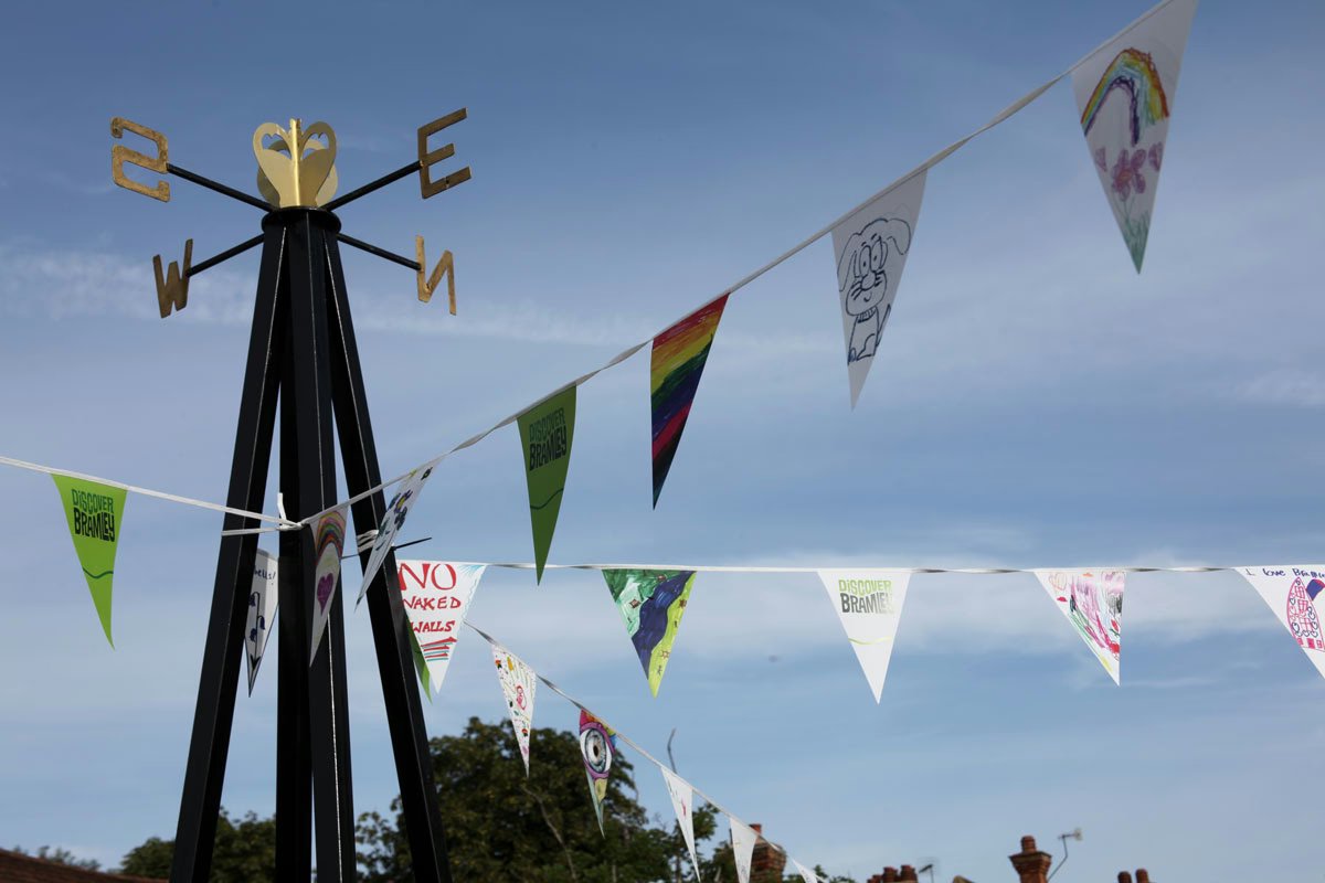 Strings of triangular bunting flags, some printed with the Discover Bramley logo and others hand-painted by children, hanging from a black weathervane pole against a blue sky