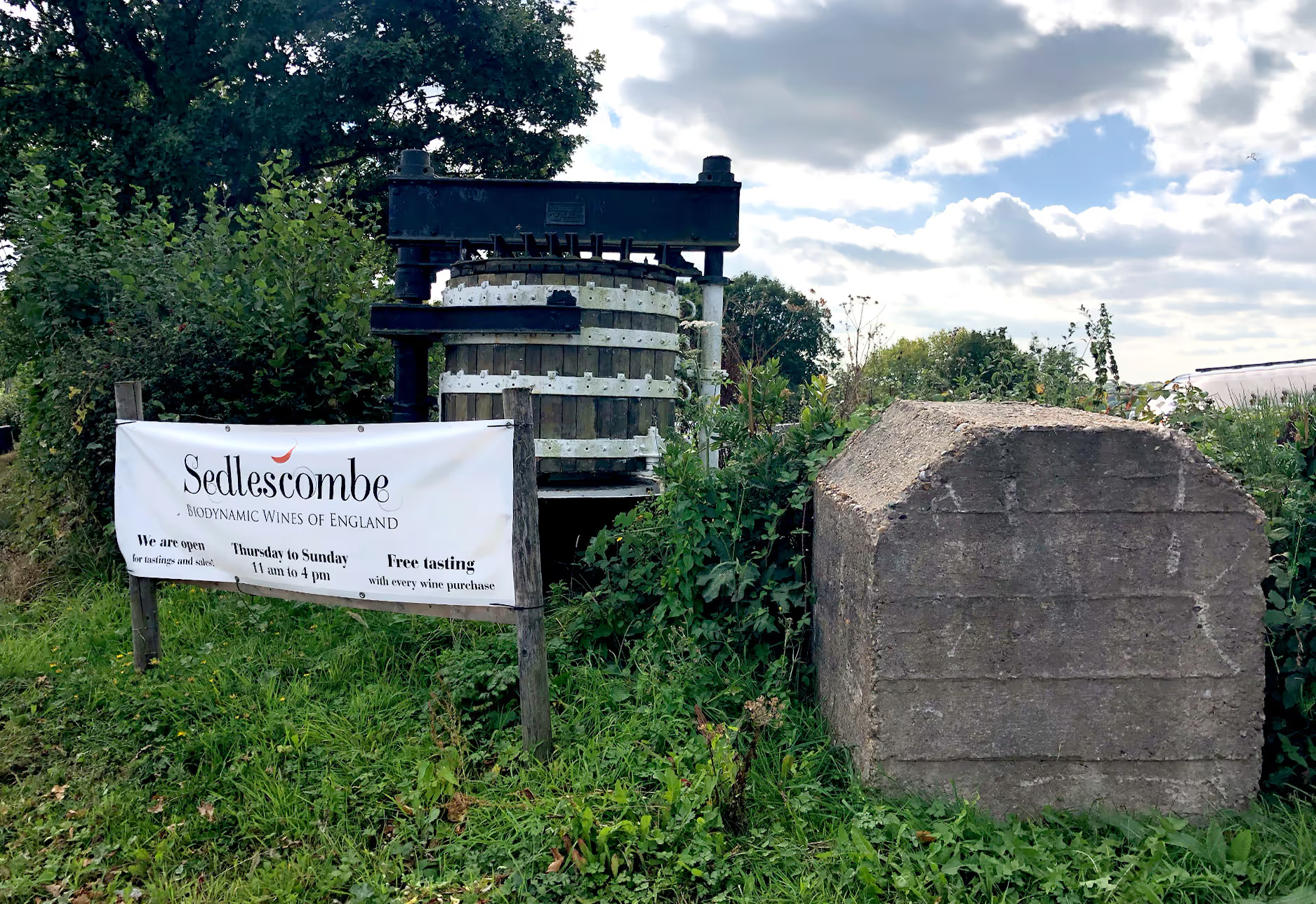A sign saying Sedlescombe next to a large decorative keg amongst foliage