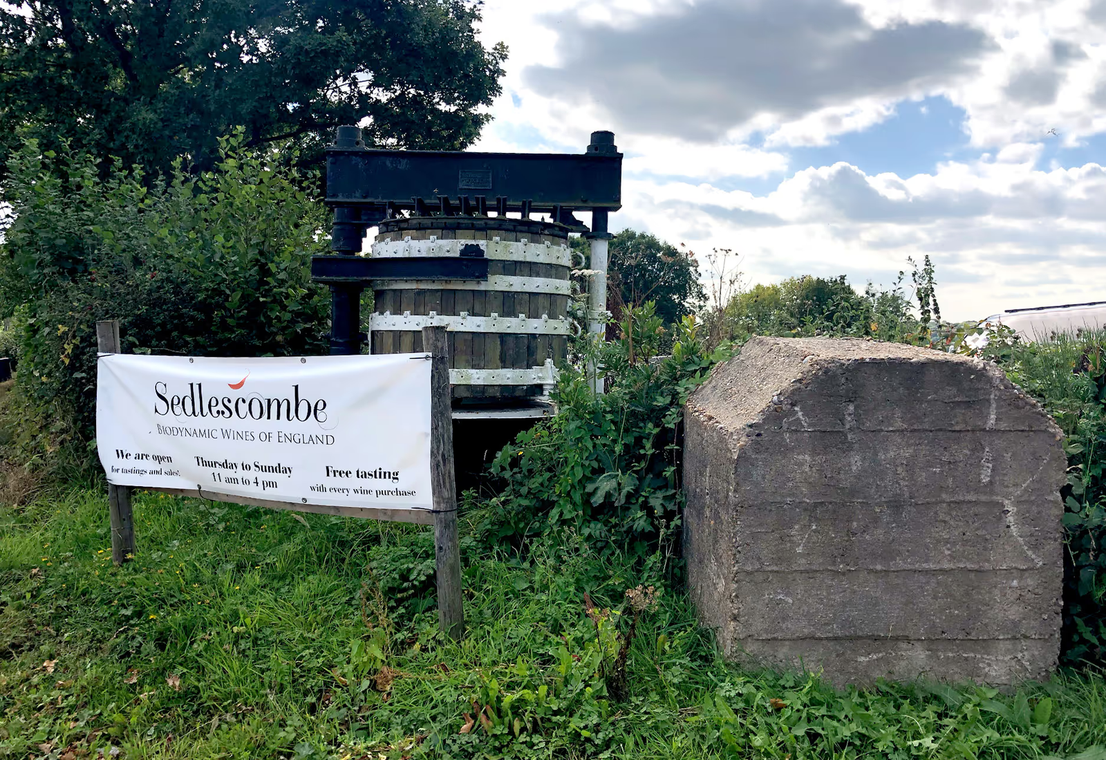 A sign saying Sedlescombe next to a large decorative keg amongst foliage