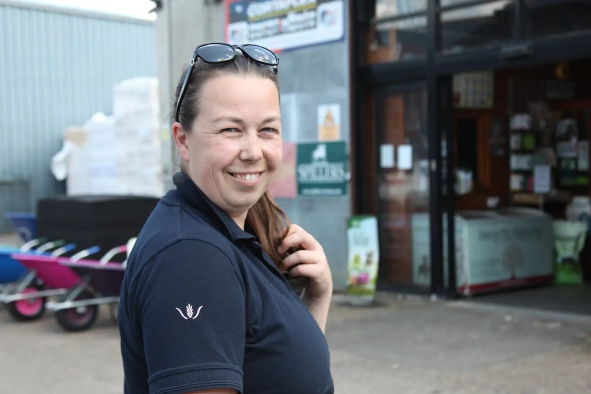 A smiling woman in a navy polo shirt stands outside the Whipley Manor Farm feed store entrance with a Spillers sign visible behind her.