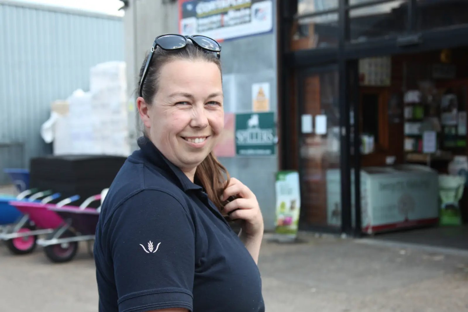 A smiling woman in a navy polo shirt stands outside the Whipley Manor Farm feed store entrance with a Spillers sign visible behind her.