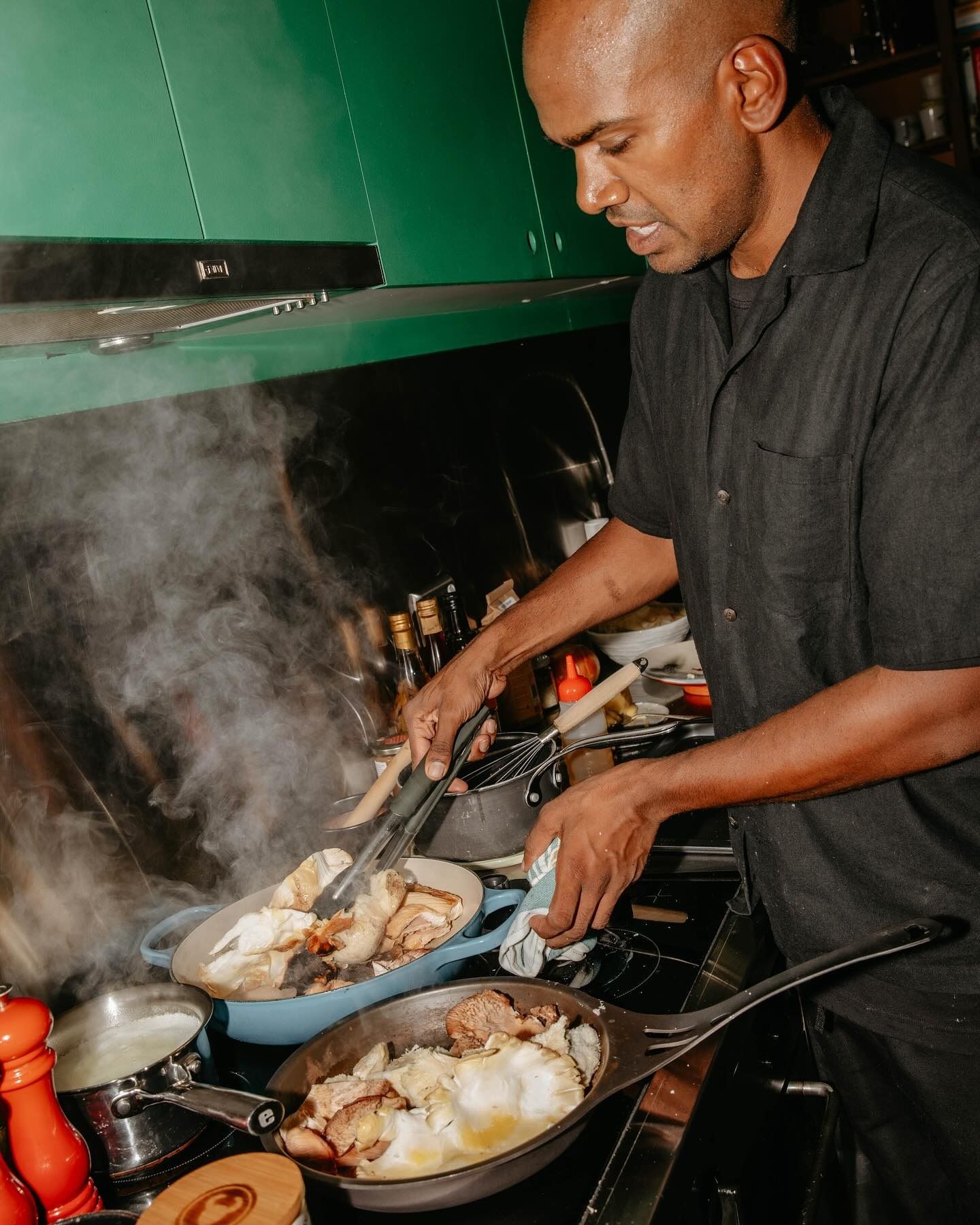 Dishan cooking mushrooms over two pans in a green kitchen, steam rising